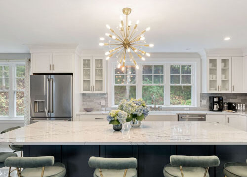 Modern kitchen with marble island, floral centerpiece, teal stools, stainless steel appliances, and large window.