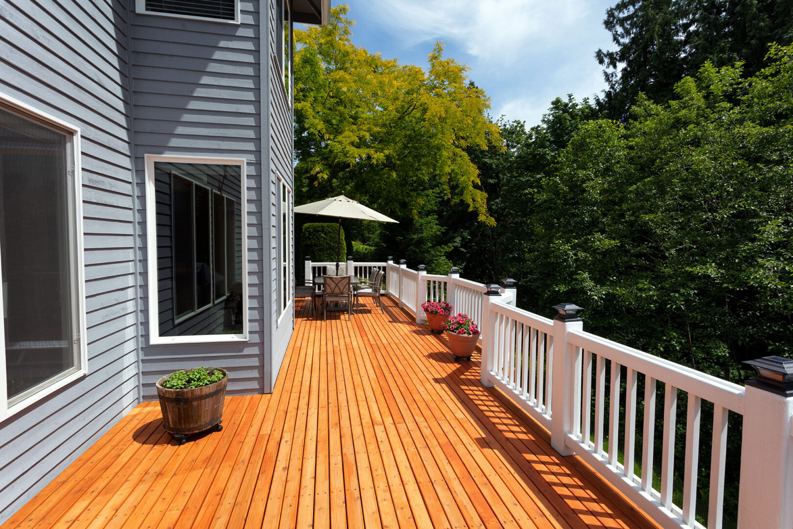 Spacious wooden deck with chairs and umbrella, lined with flower pots, next to a gray house, surrounded by lush green trees.