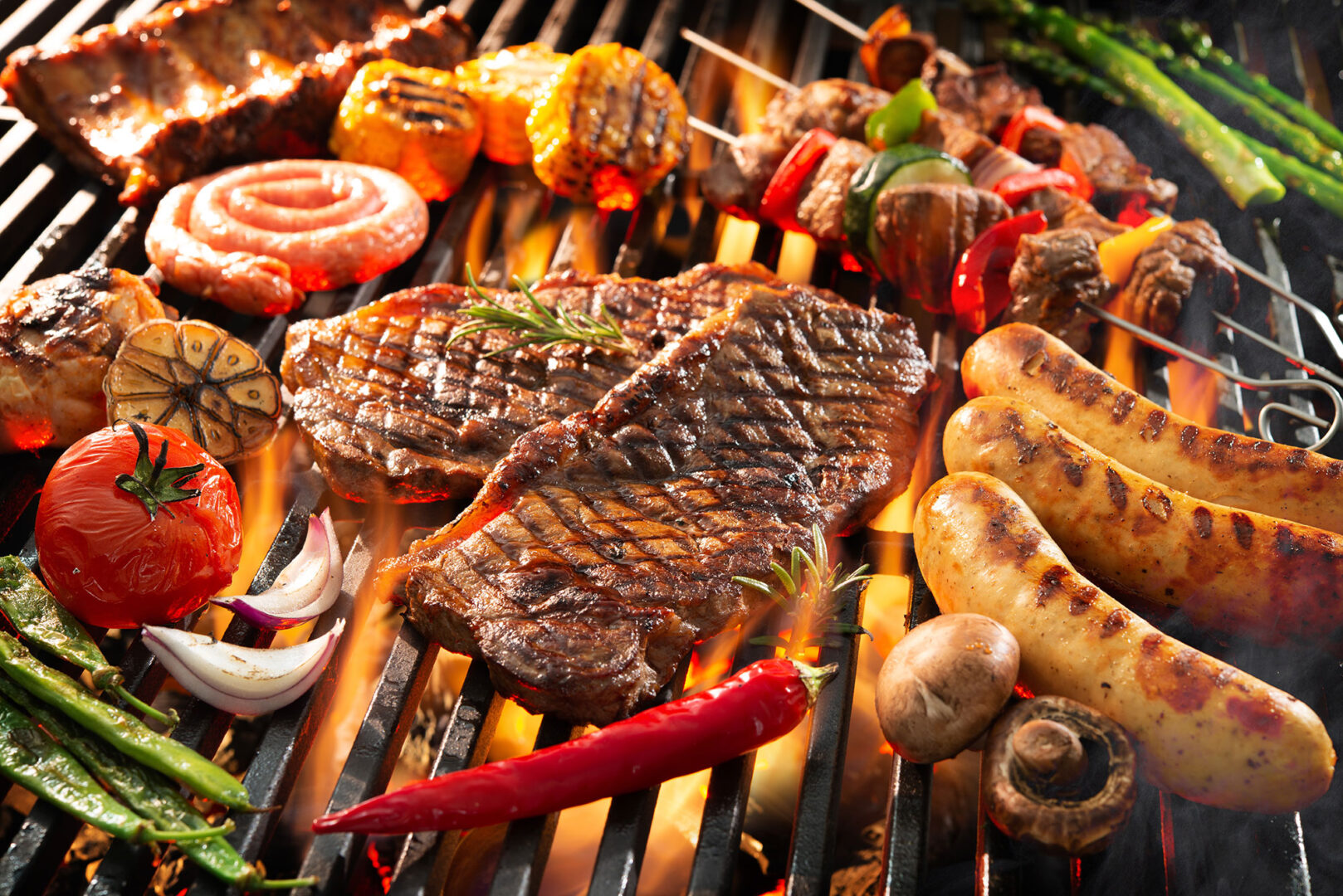 Assorted meats and vegetables grilling on barbecue, including steak, sausages, tomatoes, corn, and mushrooms.