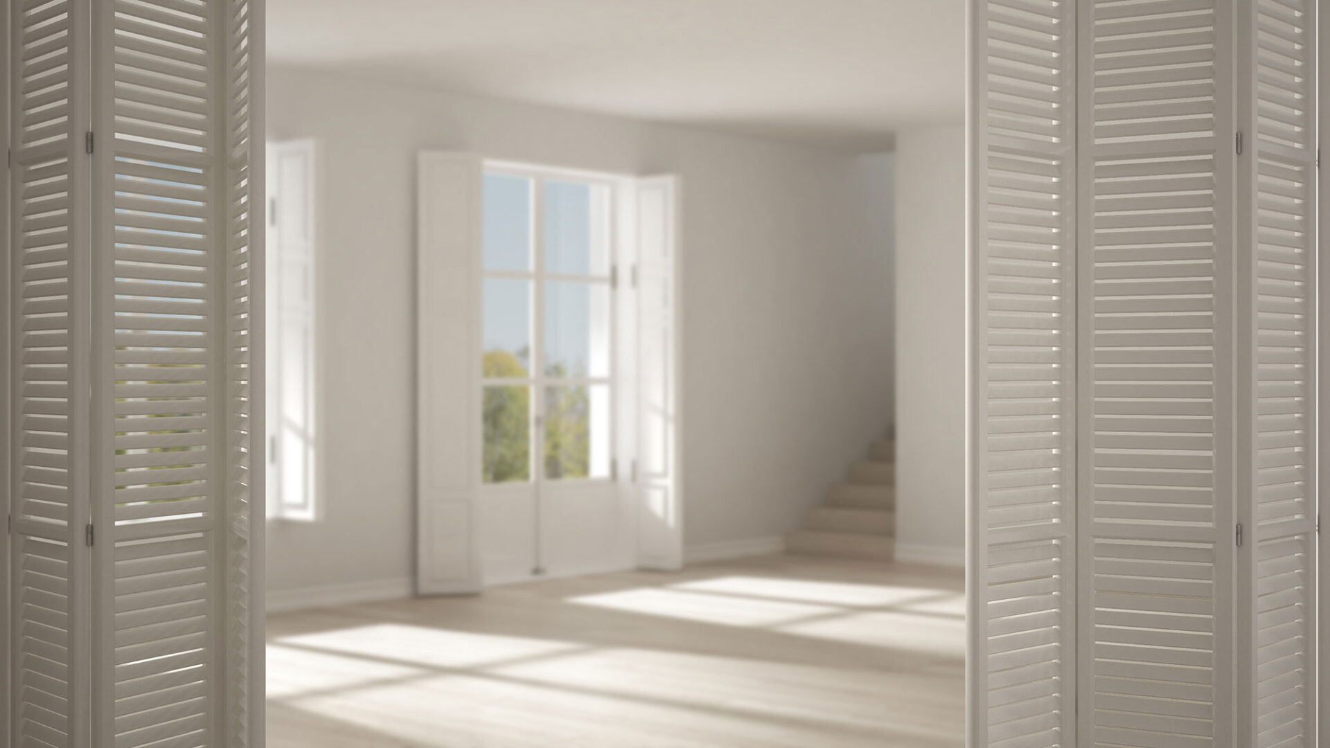 Sunlit empty room with large windows, white walls, and an open staircase in the background.