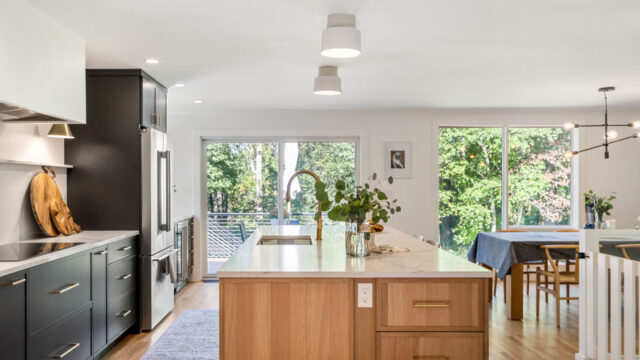 Modern kitchen with an island in the foreground, surrounded by black cabinets, and a dining area by large windows.