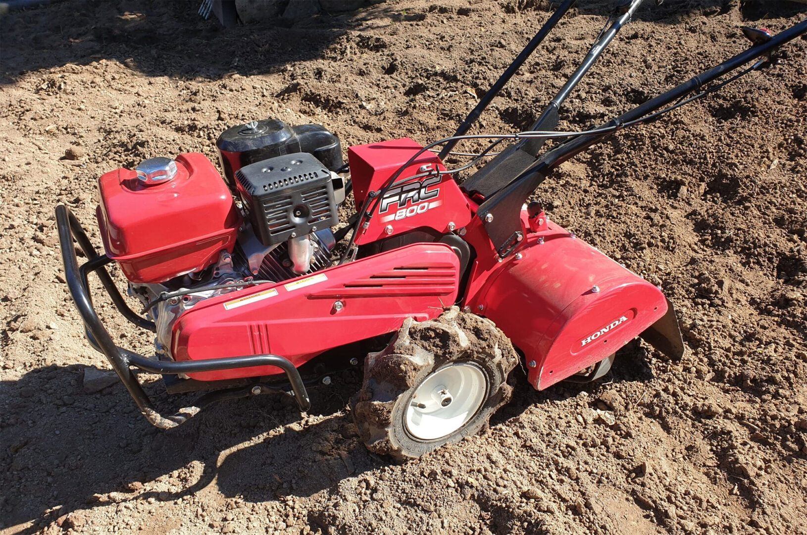 Red rotary tiller on freshly tilled soil, featuring a powerful engine and sturdy handles for gardening or landscaping.