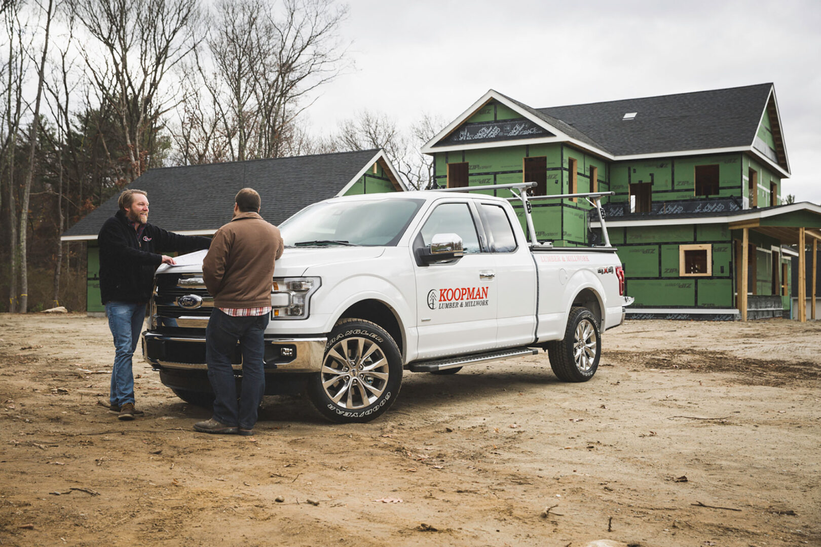 Truck parked in front of a house under construction with two people discussing plans nearby.