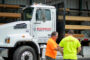 Two people in bright shirts talking beside a white truck parked in front of wooden boards at a construction site.