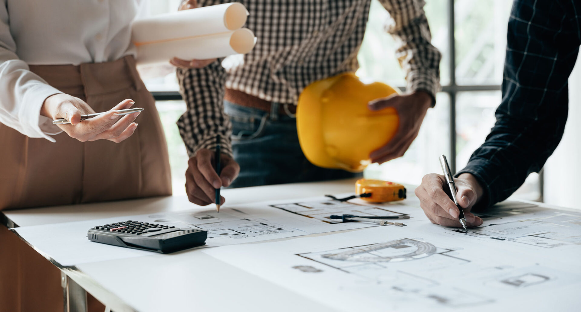 Architects reviewing construction blueprints at a desk with hard hat, measuring tape, and calculator.