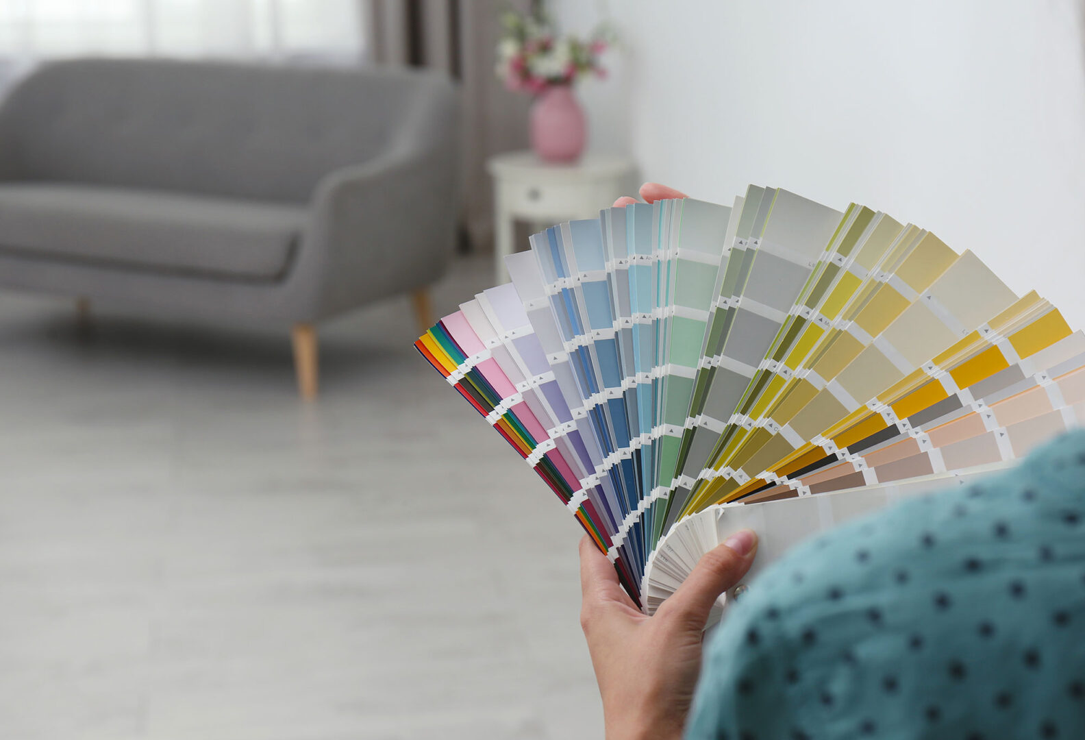 Person holding a selection of paint color swatches in a bright, modern living room with a gray sofa in the background.
