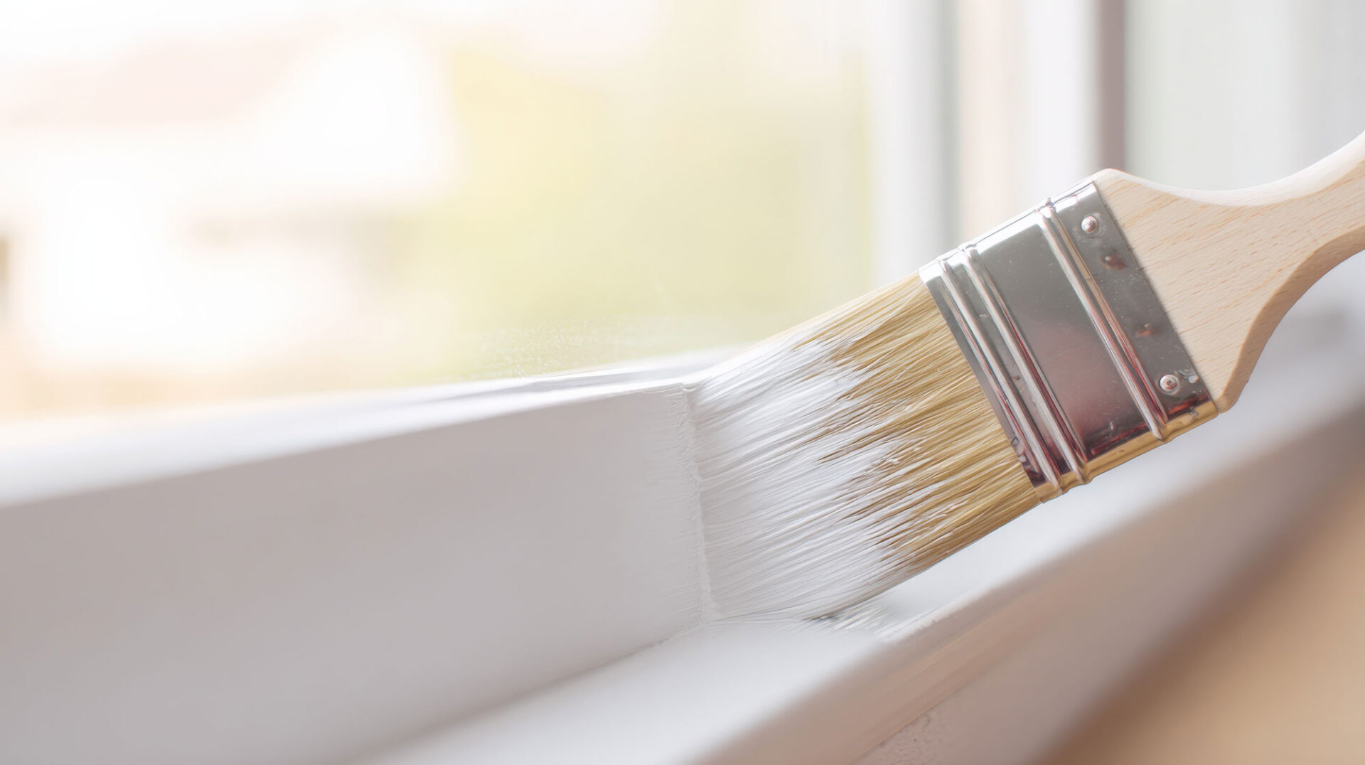 Paintbrush applying white paint on a windowsill near a sunlit window background.