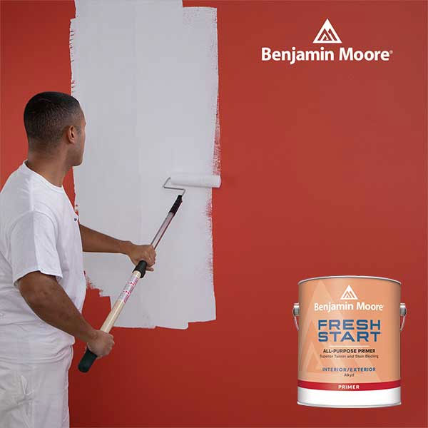 Man painting a red wall white with a roller, Benjamin Moore paint can in the foreground.