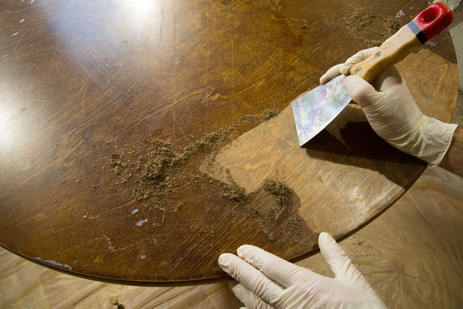 Person using a scraper to remove old finish from a wooden table surface, wearing protective gloves.