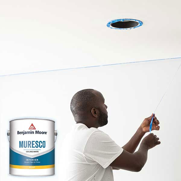 Man painting ceiling with a paint roller near a can of Benjamin Moore paint in a white room.