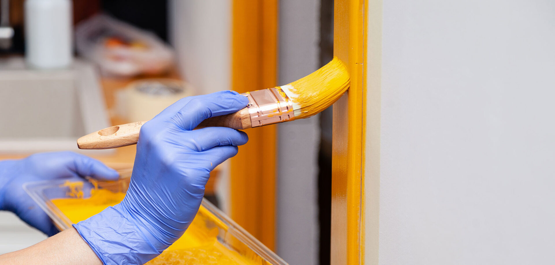 Hand in blue glove painting a door frame with yellow paint.