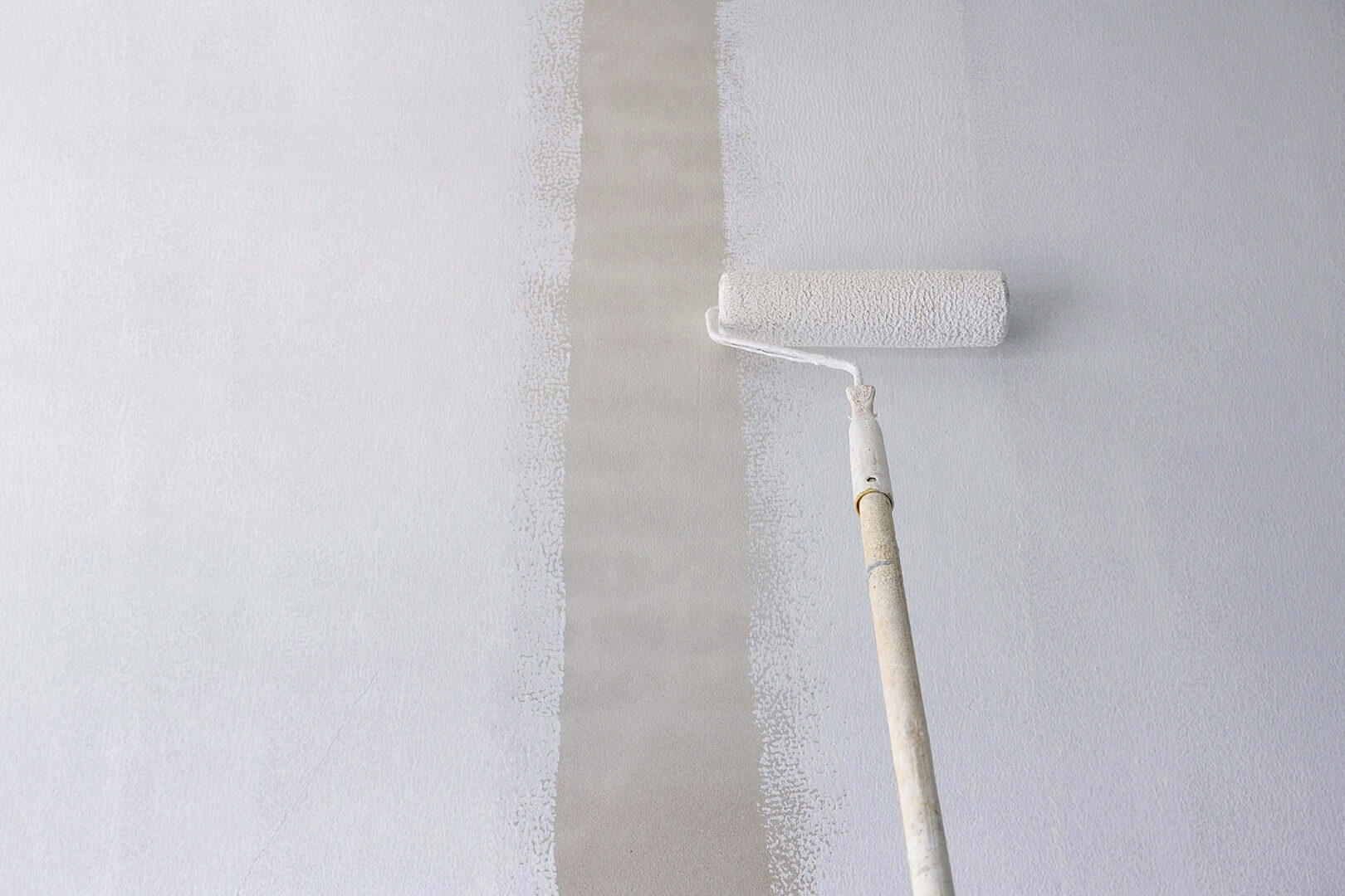 A paint roller applying a new coat of beige paint on a white wall, halfway through the process.