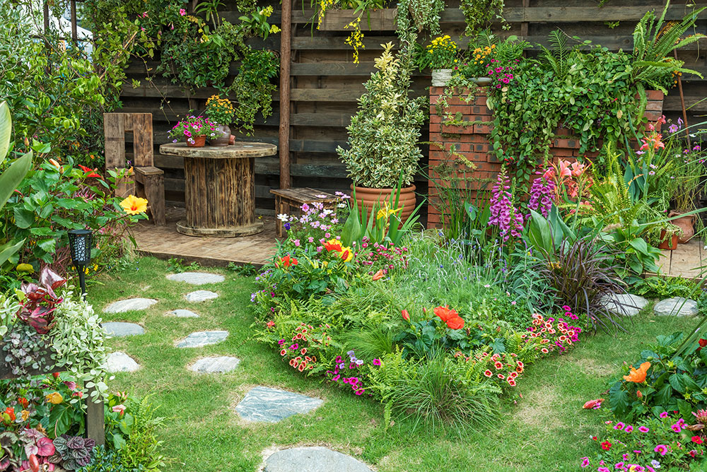 Colorful garden with lush plants, stone pathway, and wooden seating area against a wooden fence. Bright flowers in bloom.