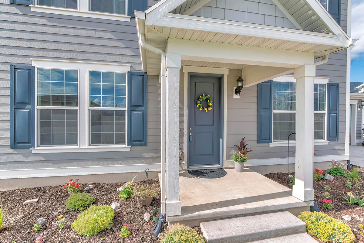 Front porch of a house with blue door, a welcome wreath, potted plant, and garden landscaping.