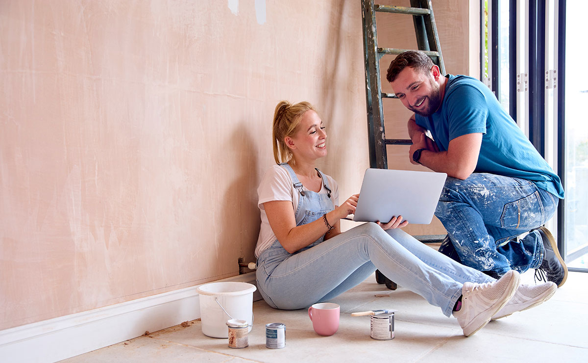 Couple in overalls sitting on floor, using a laptop, planning home renovation with paint supplies and a ladder in the background.
