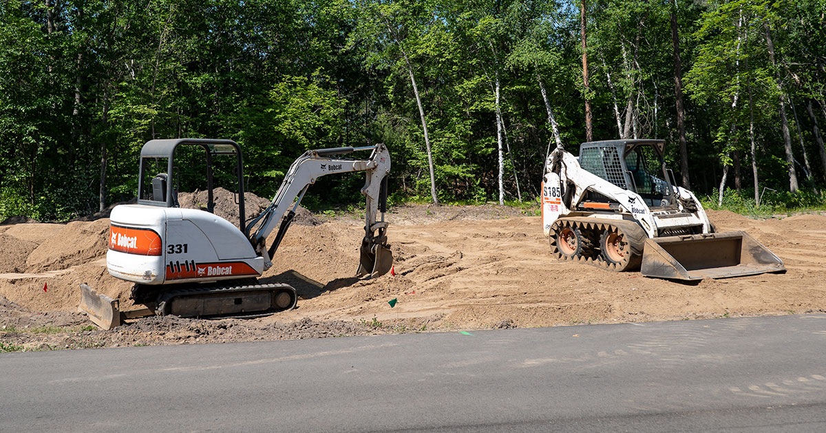 Two construction vehicles on a sandy site with trees in the background.
