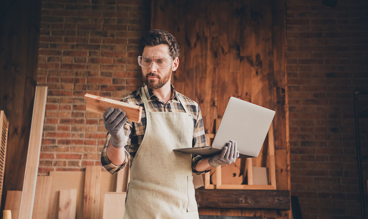 Man in apron holding wooden plank and laptop, working in wood workshop.